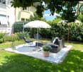 Well-maintained garden with a patio area featuring a sofa, chairs, a table, and an umbrella, surrounded by greenery and a potted plant.