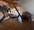 A spacious, well-lit living area with wooden flooring, a grand piano, a staircase leading to an upper level, and several seating options including a chair and small tables.