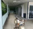 Balcony with tiled floor, scooter parked, surfboard leaning against scooter, covered area with concrete ceiling, glass railing, and outdoor furniture visible in the background.