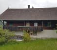 Traditional two-story wooden house with a sloped roof, green shutters, and a balcony overlooking a grassy field and forested hills.