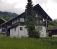 Traditional Swiss wooden house with green shutters, surrounded by lush greenery and a wind turbine in the background.