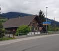 Traditional wooden house with green shutters and a sloped roof, situated near a road with a signpost and surrounding trees and mountains.