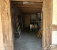 A narrow wooden-framed corridor with a concrete floor, leading to a storage area with tools and equipment.