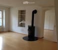 Empty room with wooden flooring, white walls, a black wood-burning stove, a built-in shelf, and a window with a radiator below it.