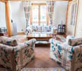 A cozy living room with a patterned sofa, two armchairs, a coffee table, side table, bookshelf, and large windows with curtains.