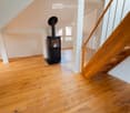 Bright hallway with wooden flooring, white walls, and a modern black wood-burning stove. A wooden staircase with a metal railing leads upwards.