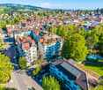 Aerial view of a residential area with multiple buildings, red-tiled roofs, green spaces, and a swimming pool in the foreground.
