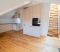 Modern kitchen with light-colored cabinets, wooden flooring, and a small staircase leading upwards.