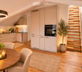 Modern kitchen and dining area with light-colored cabinets, a round wooden dining table, four chairs, a pendant light, and a staircase leading upwards.