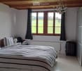 Cozy bedroom with a large bed, nightstand, lamp, and suitcase near the window. The room features wooden beams on the ceiling and large windows with dark curtains, offering a view of a green field.