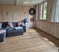 Cozy living room with wooden flooring, grey sectional sofa with pink and grey pillows, dartboard on wall, storage unit, and large windows with curtains.