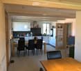 Open-plan living and dining area with a wooden dining table, four black chairs, a kitchen area with white cabinets, a refrigerator, and various kitchen appliances.