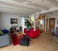 Open-plan living and dining area with wooden flooring, exposed wooden beams, a grey sofa, red sofa, coffee table, ottoman, dining table with chairs, bookshelf, and a staircase leading to an upper level.