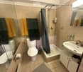 A small bathroom with beige tiled walls and floor, featuring a toilet, bathtub with a shower curtain, a sink with a mirror, and a towel rack with towels.