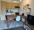 A modern, open-plan kitchen and dining area with light wooden cabinets, a central dining table with chairs, and a television on a stand. The space features a hanging pendant light and a potted plant on the counter.