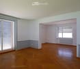 Completely empty room with wooden herringbone flooring, white walls, and large windows with shutters. No furniture or equipment.
