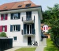 white facade, red roofs, red and black shutters, balconies, garage, green grass, trees, car parked