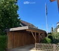 Wooden garage with a slanted roof, surrounded by greenery and a wooden fence.