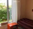 Bright bedroom with a bed, nightstand, lamp, and books. Large window with white curtains offers a view of greenery outside.