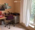 A well-organized home office with a wooden desk, purple office chair, printer, and various office supplies. The room features framed artwork on the walls and a large window with white curtains.