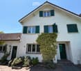 Two-story white house with green shutters, ivy-covered walls, and a tiled roof. Features include a front door, multiple windows, and a small garden area with shrubs.