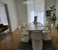 Bright room with wooden flooring, large windows with sheer curtains, a sofa, a dining table with chairs, a laptop, a potted plant, and a picture frame on the wall.