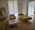 Living room with wooden flooring, large windows with white curtains, a sofa, a chair, a coffee table with a chessboard, and a small table with chairs.