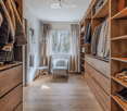A well-organized walk-in closet with wooden shelves and drawers, a small seating area featuring a grey armchair and a round stool, and a coat rack with jackets and bags.
