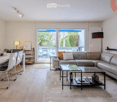 Modern living and dining area with a grey sectional sofa, glass coffee table, white dining table with chairs, and large windows opening to a balcony.