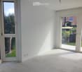 Empty room with white walls, tiled floor, and large windows offering views of greenery and a neighboring house.