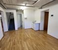 Empty room with wooden flooring, white walls, and several open doors leading to other areas, including a staircase and a small kitchenette area.
