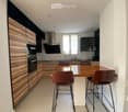 Modern kitchen with wooden cabinets, stainless steel appliances, a central island with a wooden table and four brown chairs, and a window with white blinds.