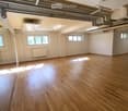 Empty room with wooden flooring, white walls, exposed ceiling pipes, and several windows allowing natural light.