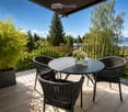 Balcony with a round table, four wicker chairs, and a vase of white flowers, overlooking a scenic view of trees and a body of water.