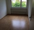 Empty room with wooden flooring, white walls, large window with view of greenery, radiator below window, and door on right side.
