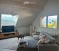Modern living room with large windows offering a scenic mountain view, featuring a grey sectional sofa, coffee table, TV, guitar, and decorative elements.