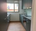 Modern kitchen with dark grey cabinets, light wooden flooring, and a window offering a view of the balcony and surrounding buildings.