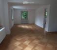 Empty room with wooden herringbone flooring, white walls, and large windows allowing natural light. Radiators are visible along the walls.
