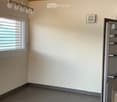 Empty room with wooden ceiling beams, grey tiled floor, large window with blinds, and a partially visible kitchen area with a stainless steel appliance.