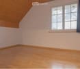 Empty room with wooden flooring and white walls, featuring a large window with dark curtains and a slanted wooden ceiling typical of an attic space.