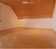 Empty room with wooden flooring and white walls, featuring a slanted wooden ceiling and a window with multiple panes.