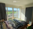 Bright bedroom with large windows offering a scenic view, featuring a bed with a colorful blanket, a nightstand, a wardrobe, a clothing rack, and a chair.