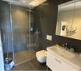 Modern bathroom with dark tiled walls, a glass-enclosed shower, a white sink with a mirror cabinet, a toilet, and stacked washing machine and dryer units.