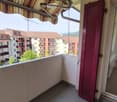 Balcony with grey concrete floor, red shutters, striped awning, and view of neighboring buildings and greenery.