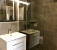 Modern bathroom with a white sink, mirror, and glass-enclosed shower area. The walls are tiled in a dark, textured pattern, and the fixtures are sleek and contemporary.
