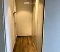 A narrow hallway with light wooden flooring, white walls, and recessed ceiling lights. The hallway features a closed door on the left and a mirrored wall on the right.