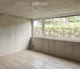 Minimalist unfurnished room with exposed wooden ceiling, concrete walls, and large window overlooking a landscaped garden.