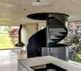 Modern hallway with a sleek black spiral staircase, wooden ceiling, and large windows overlooking a green landscape.