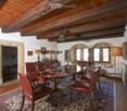 Elegant dining room with exposed wooden beams, stone walls, and large windows. Features a round wooden dining table surrounded by red leather chairs, lamps, and artwork.
