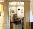 A small, cozy room with stone walls and a tiled floor, featuring a patterned armchair, a small wooden table, a wooden chair, and framed artwork on the walls.